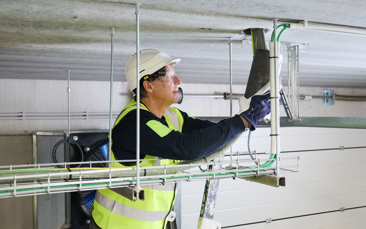 Montážny pracovník na rebríku pripevňuje na strop protipožiarnu manžetu, ktorá je súčasťou tesniaceho systému pre požiarne prestupy PROMASTOP®-IM CBox 125.
