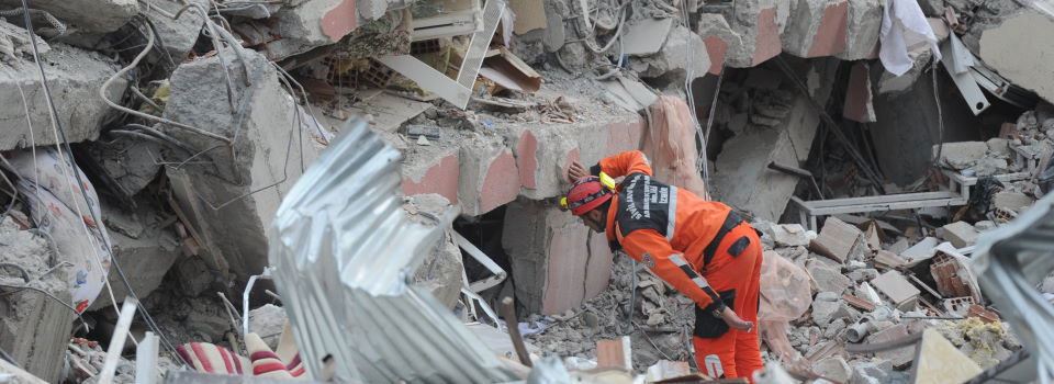 An image of a black member of a rescue team dressed in an orange suit peering into concrete rubble.