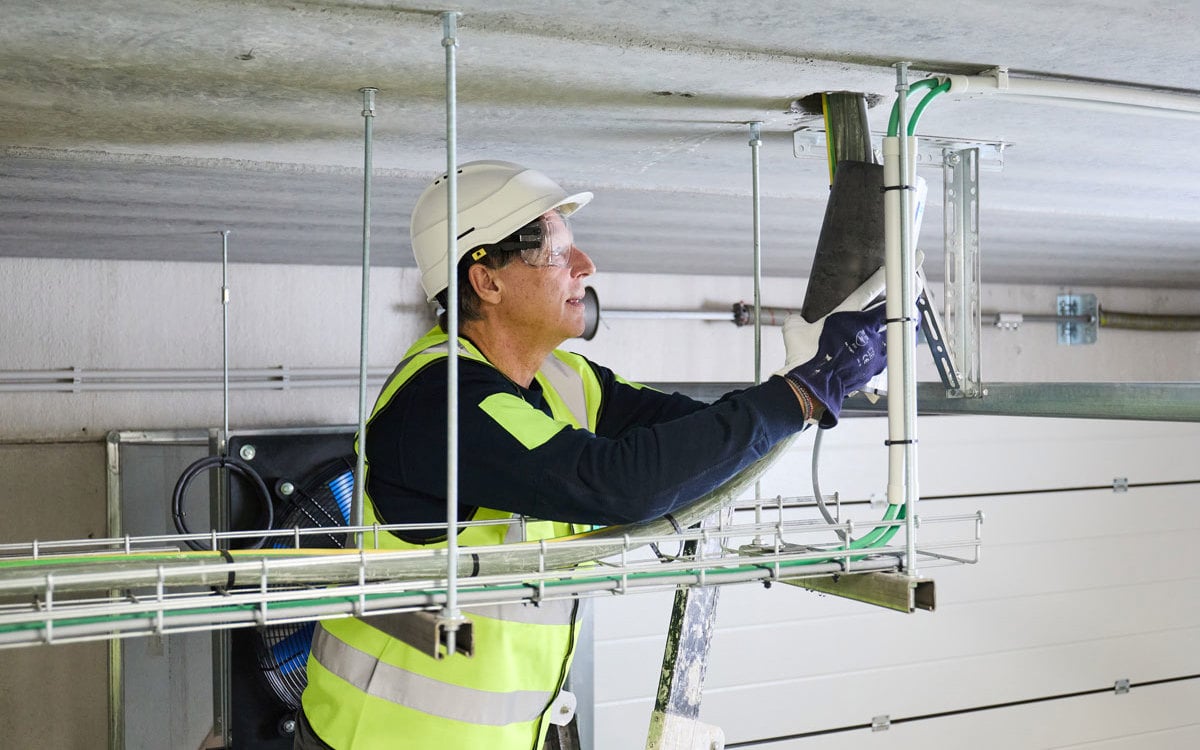 Čovek na merdevinama koji ugrađuje protivpožarnu obujmicuna plafonu gde se iza njega nalazi industrijski ventilator