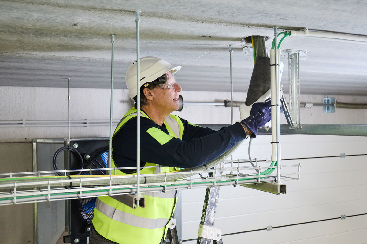 An installer on a ladder fitting a fire collar, a part of PROMASTOP®-IM CBox 125 intumescent sealing system, onto the ceiling.