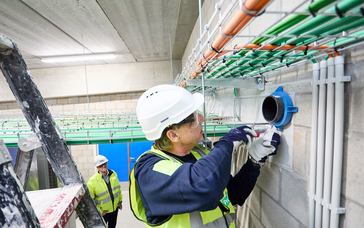 A worker in a cellar installing a firestopping solution. There is a ladder to the left and a man watching in the background.