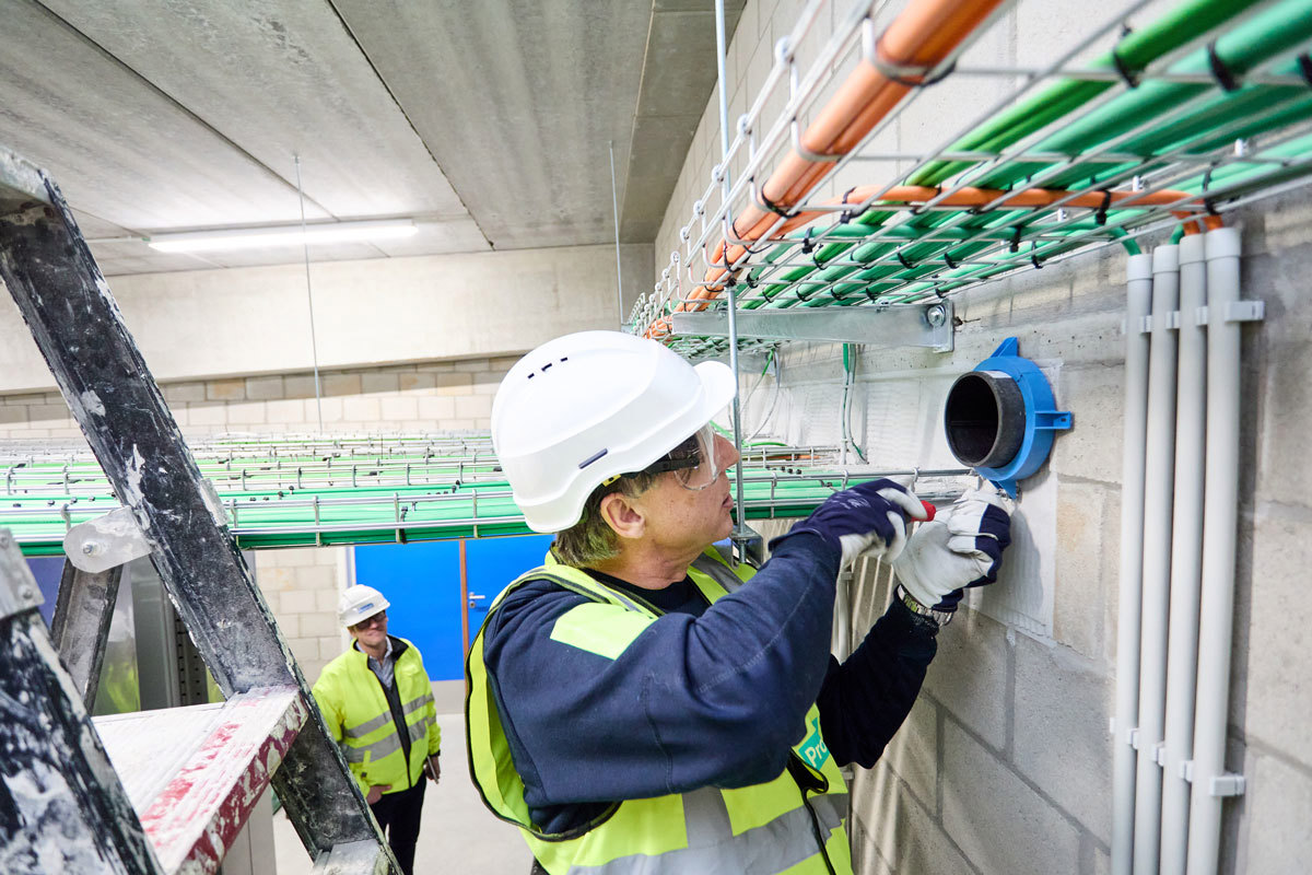 A worker in a cellar installing a firestopping solution. There is a ladder to the left and a man watching in the background.