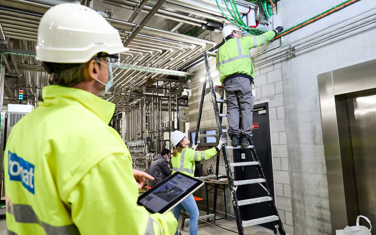 Un bărbat cu o tabletă care se uită la 3 instalatori ce instalează sigilii de penetrare folosind vopseluri și agenți de etanșare PROMAT.