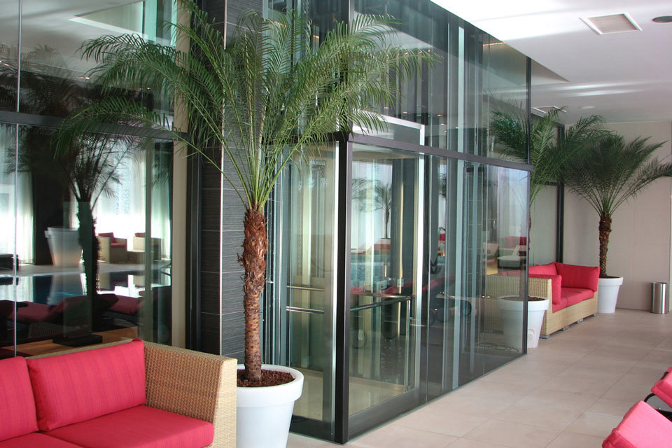 Prehodna SR požarna vrata in požarna zasteklitev brez profilov Promat®-SYSTEMGLAS 30 v notranjosti hotela Kempinski Palace v Portorožu, levo in desno od zastekljenih vrat je sedežna garnitura in okrasne manjše zelene palme. 