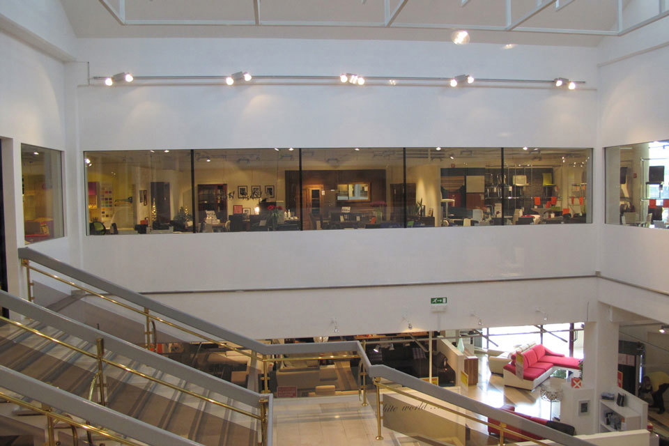 Pogled na bel razstavni prostor in požarno odporno steklo brez okvirjev Promat®-SYSTEMGLAS F1 60. Steklena okna so po celotnem obsegu prostora v Pohištvenem centru Lesnina v Kopru, v levem delu slike so stopnice, ki vodijo navzdol. 