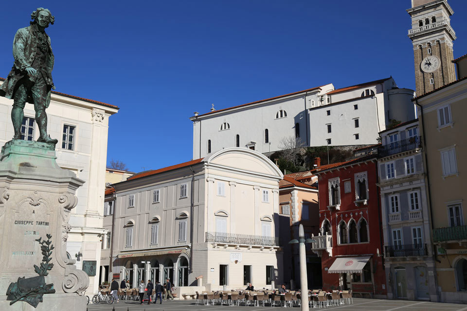 Pogled iz Tartinijevega trga, levo je spomenik Tartiniju, na sredini je bela zunanjost Mestne galerije Pirana, desno so rdeča, bela in rumena hiša na trgu. 