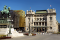 Mogočna rumeno rjava steklena zunanjost hotela Courtyard Marriott Beograd na levi strani slike, desno je mogočna bela stavba, v ospredju pa je spomenik moža na konju. 
