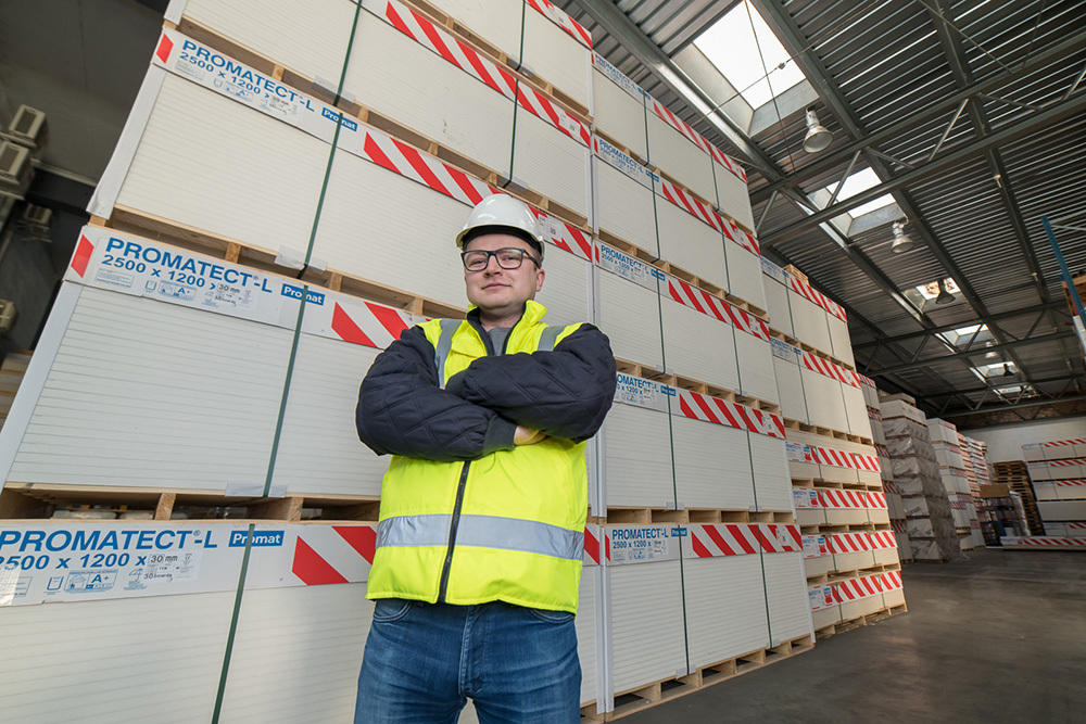 Slika Kamila Ziółkowskega v skladišču s paletami plošč Promatect-L v ozadju.