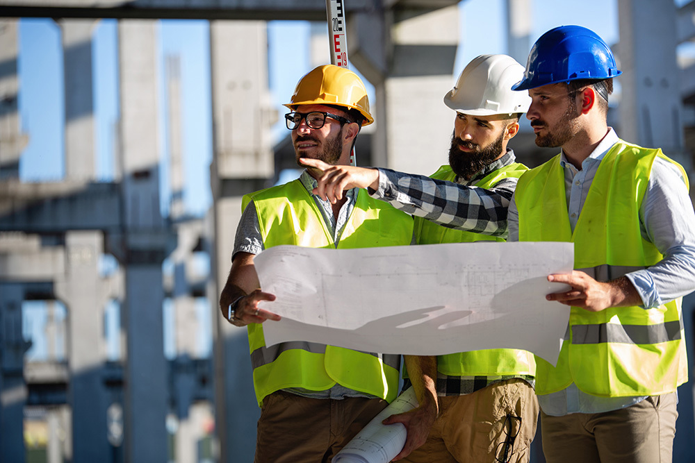 Fotografija sa tri građevinska inžinjera koji nose zaštitne šlemove, markere i pritom  razgovaraju o tehničkom planu gledajući u plan projekata.