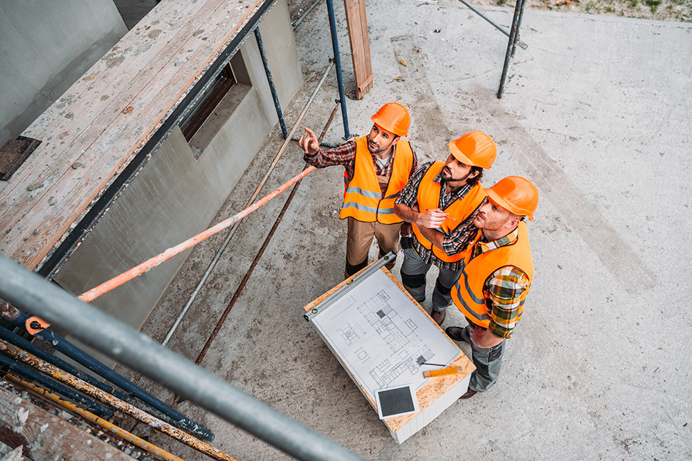 Fotografija trojice građevinskih inžinjera koji nose zaštitnu opremu, razgovaraju o planu prodora instalacija koji je ispred njih i gledaju u projekat.