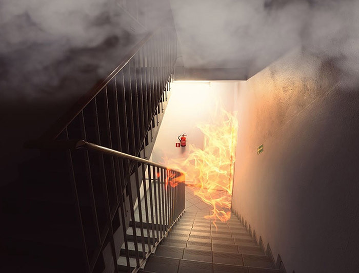 Zdjęcie. Schody w budynku w dół. Po lewej stronie poręcz. Góra kadru jest wypełniona dymem. Na dole, z pomieszczenia po prawej stronie wydobywają się płomienie, dalsze przejście w dół jest zablokowane przez płomienie. Na ścianie za płomieniami wisi czerwona gaśnica.