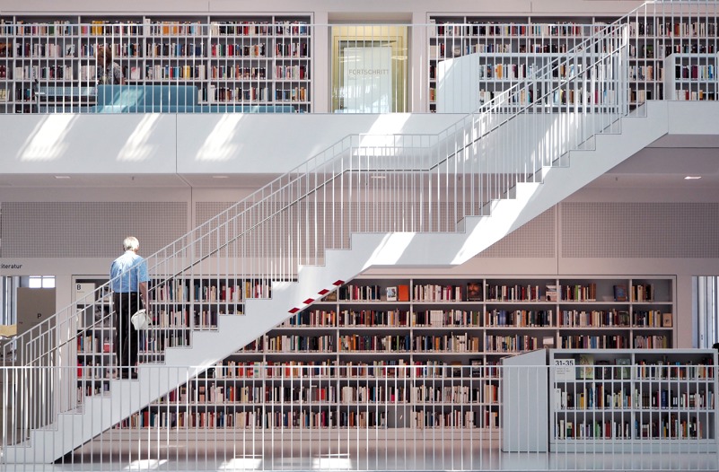Notranjost knjižnice z belo konstrukcijo in belimi stopnicami, ki gredo diagonalno v zgornje nadstropje, na njih stoji oseba. V ozadju so knjige. 