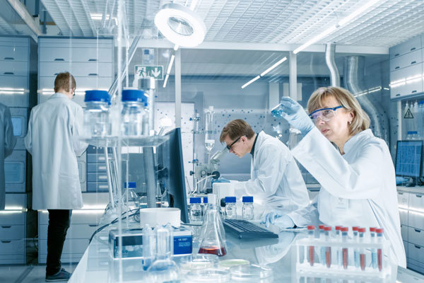 Štirje raziskovalci v belih haljah v svetlem raziskovalnem laboratoriju preučujejo in gledajo v aparature, v ospredju je veliko epruvet. 