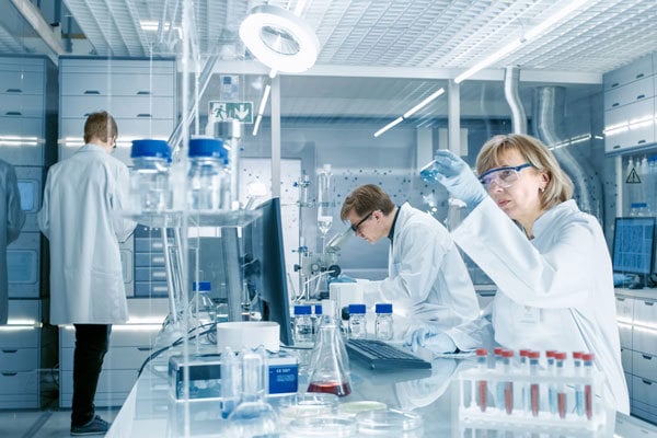 Štirje raziskovalci v belih haljah v svetlem raziskovalnem laboratoriju preučujejo in gledajo v aparature, v ospredju je veliko epruvet.