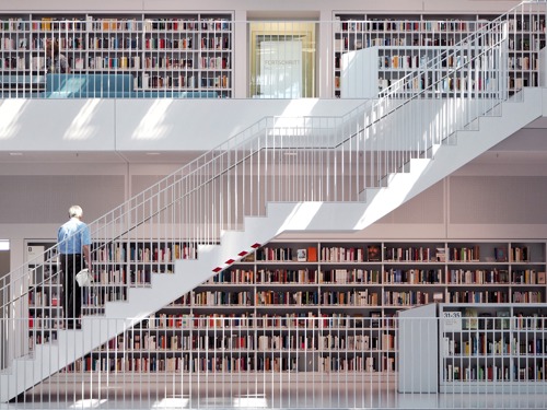Čovjek stoji na bijelim stepenicama koje vode na drugi kat moderne knjižnice i gleda u veliku zbriku knjiga.