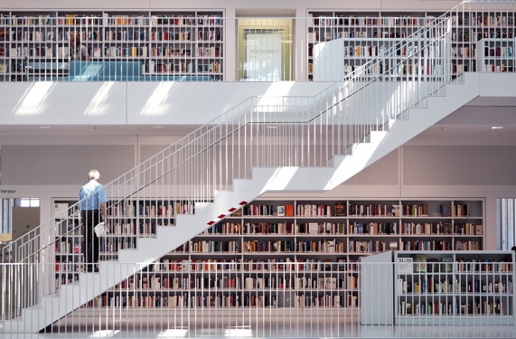 Čovjek stoji na bijelim stepenicama koje vode na drugi kat moderne knjižnice i gleda u veliku zbriku knjiga.