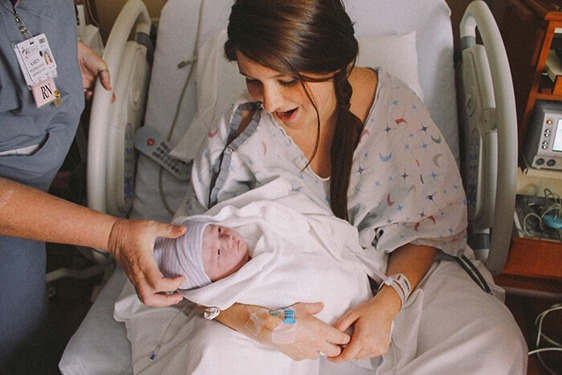 Mama drži uvijenu bebu u naručju u bolničkom krevetu dok joj bolničar u plavoj uniformi pomaže pridržavajući bebinu glavu.