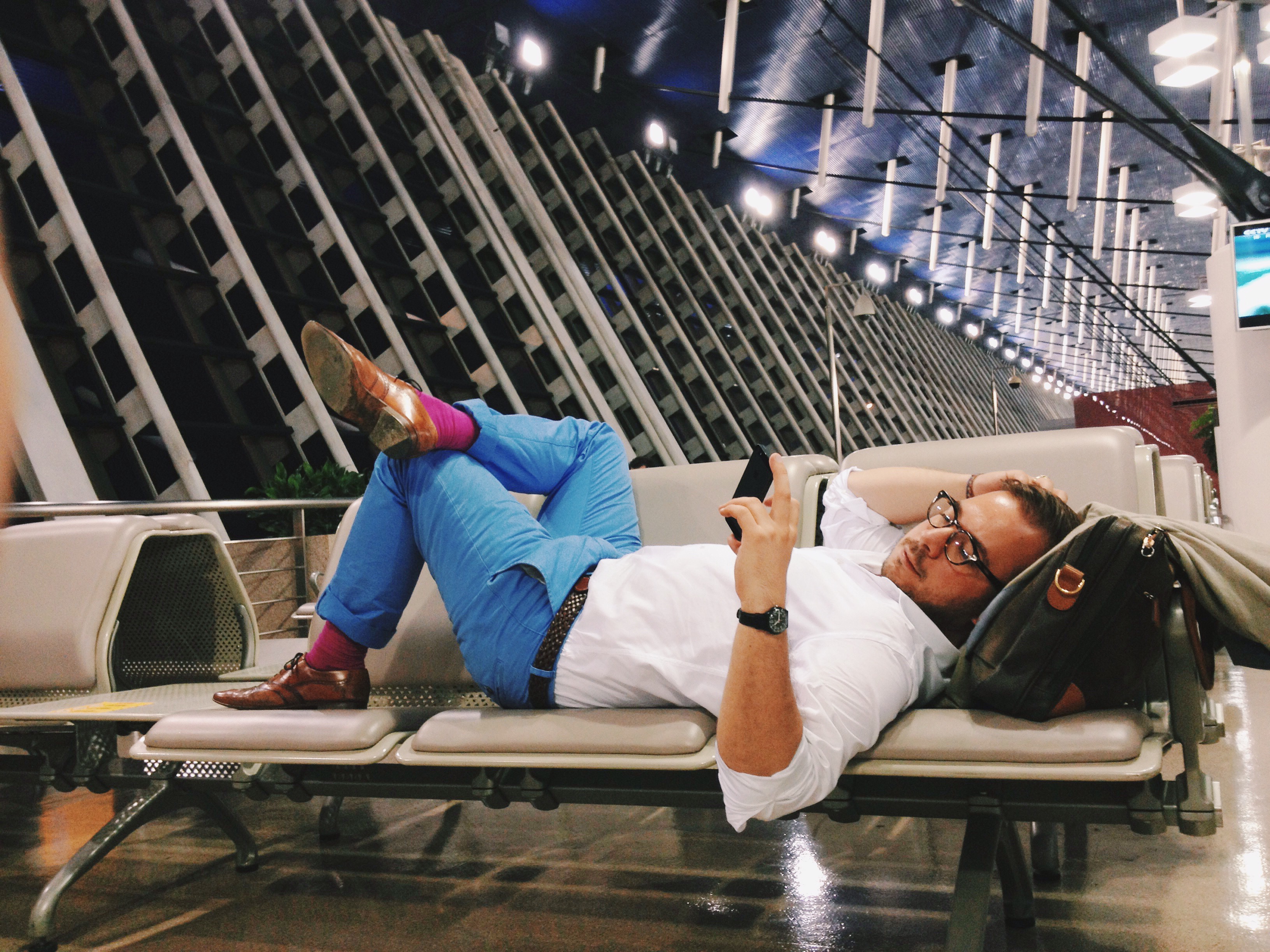 Muž leží na sedačkách v odletovej hale opretý o aktovku a pozerá sa do mobilu