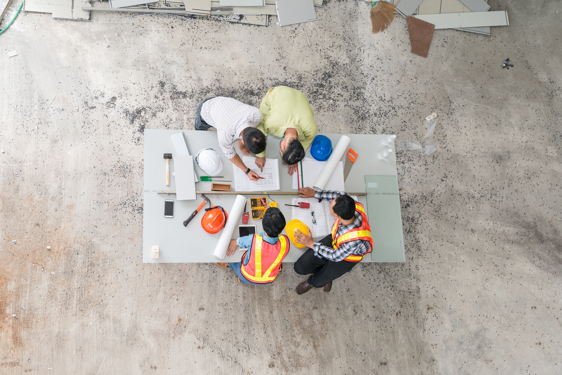 Slika s pogledom od zgoraj na mizo s tehničnimi pripomočki, za katero so štirje inženirji, ki preučujejo in rišejo načrte. 