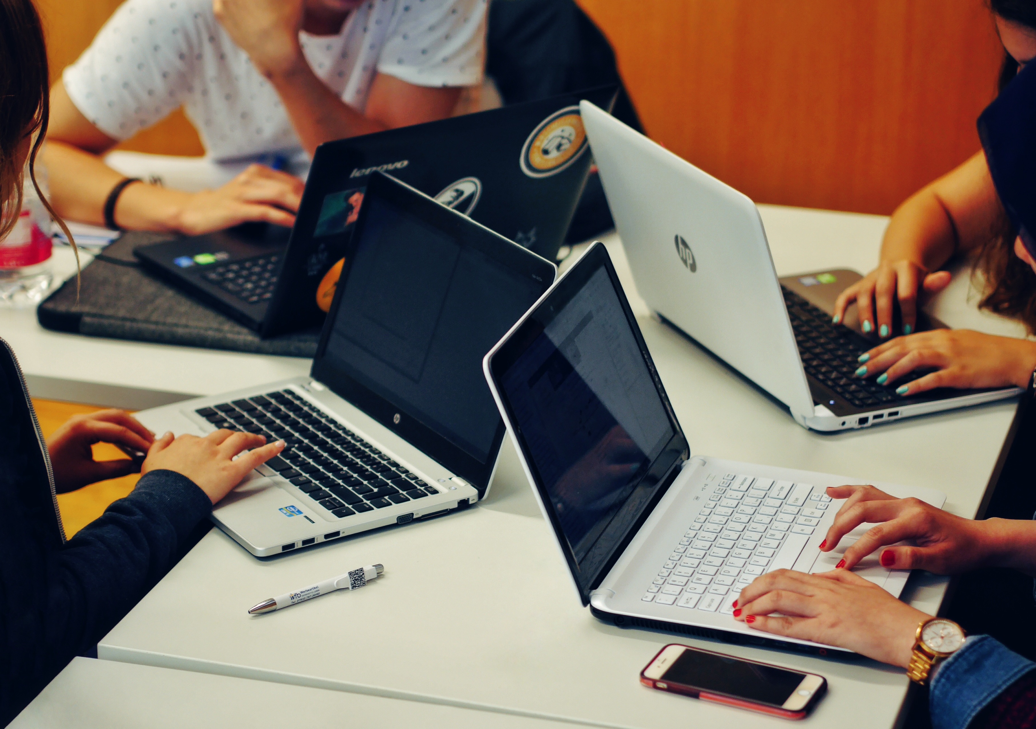 Na mizi v pisarni so postavljeni štirje črno beli prenosni računalniki, na vsakega tipka en par ženskih rok, v ospredju pa ležita prenosni telefon in pisalo.