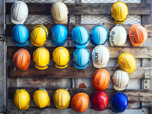 Na lesenih deskah so položene delavske čelade modrih, rumenih, oranžnih in belih barv ter ena rdeča čelada. 