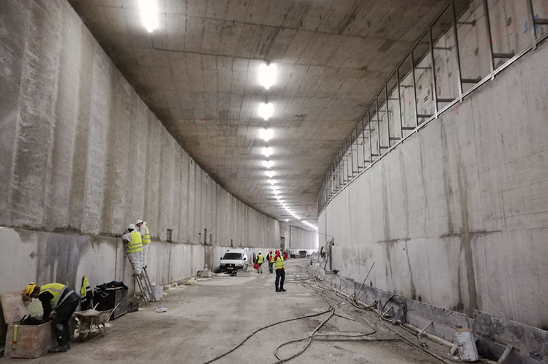 Građevinski radovi u osvetljenom betonskom tunelu sa visokom tavanicom koji skreće udesno u kom je nekoliko radnika i vozilo 