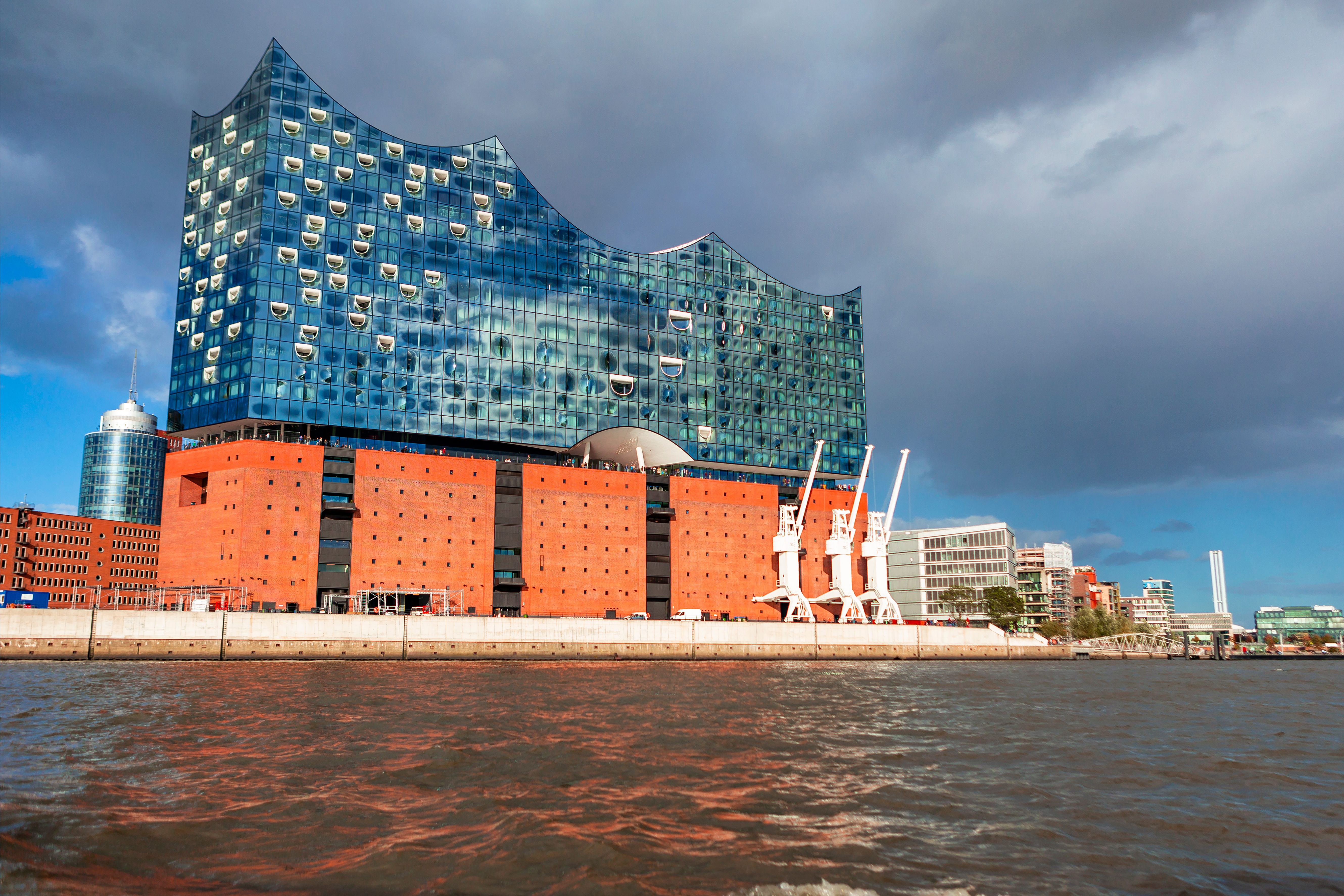 Na slici je moderna zgrada smještena je na obali rijeke. Donji dio zgrade je crvene boje, a gornji dio je u staklu sa valovitim krovom. U pozadini zgrade je grad.