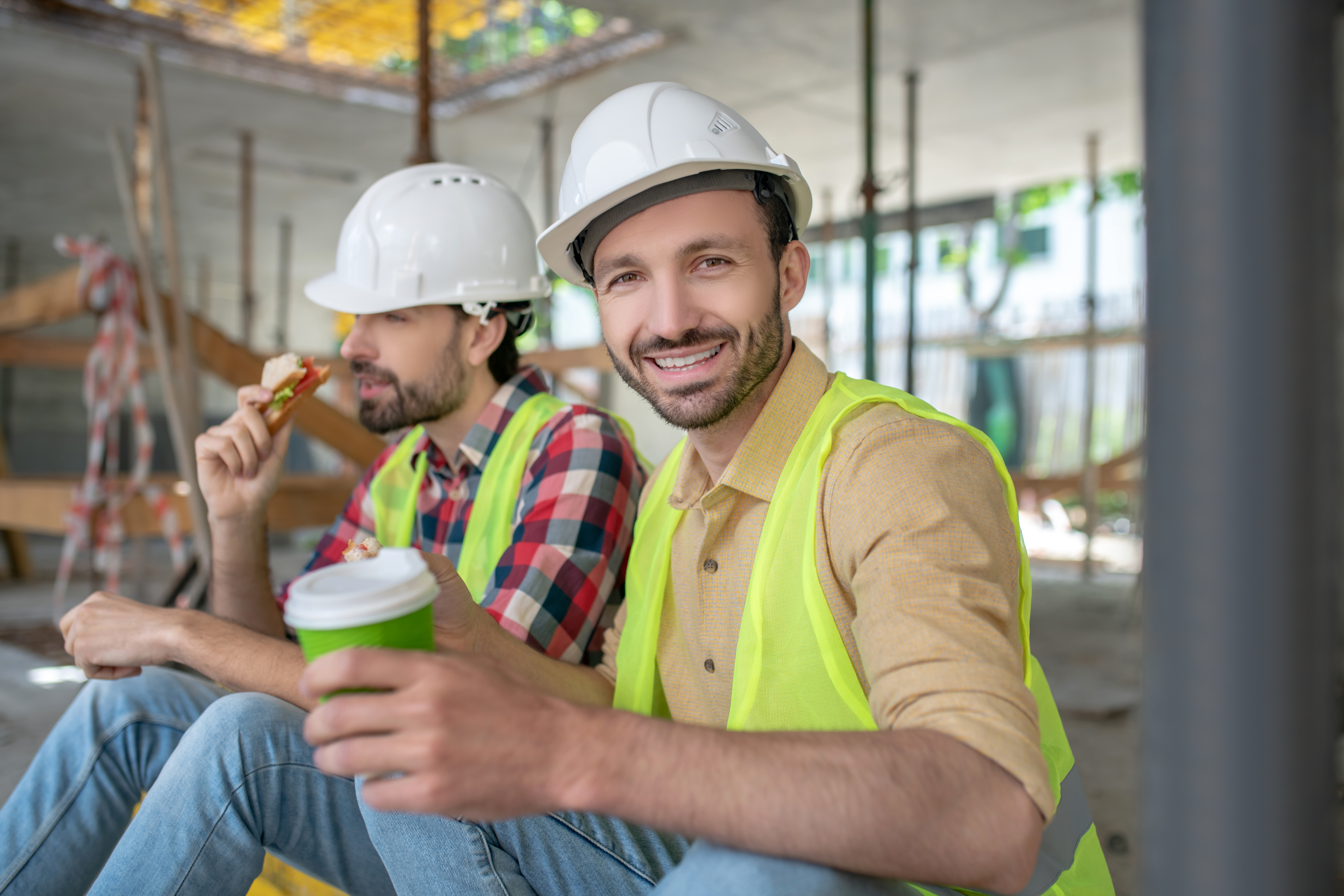 Dobro raspoloženi radnici u belim šlemovima i fluorescentnim prslucima prave pauzu za užinu u toku rada na građevini.