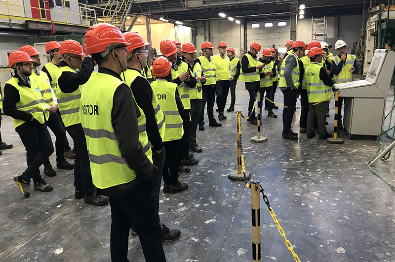 Velika skupina inženirjev v rumenih odbojnih brezrokavnikih s črnimi rokavi in rdečimi čeladami na glavi, stoji in posluša razlago v industrijskem prostoru. 