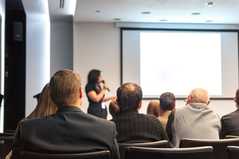 Slika iz zadnjih redova konferencijske dvorane gdje žena drži predavanje a publika sluša.