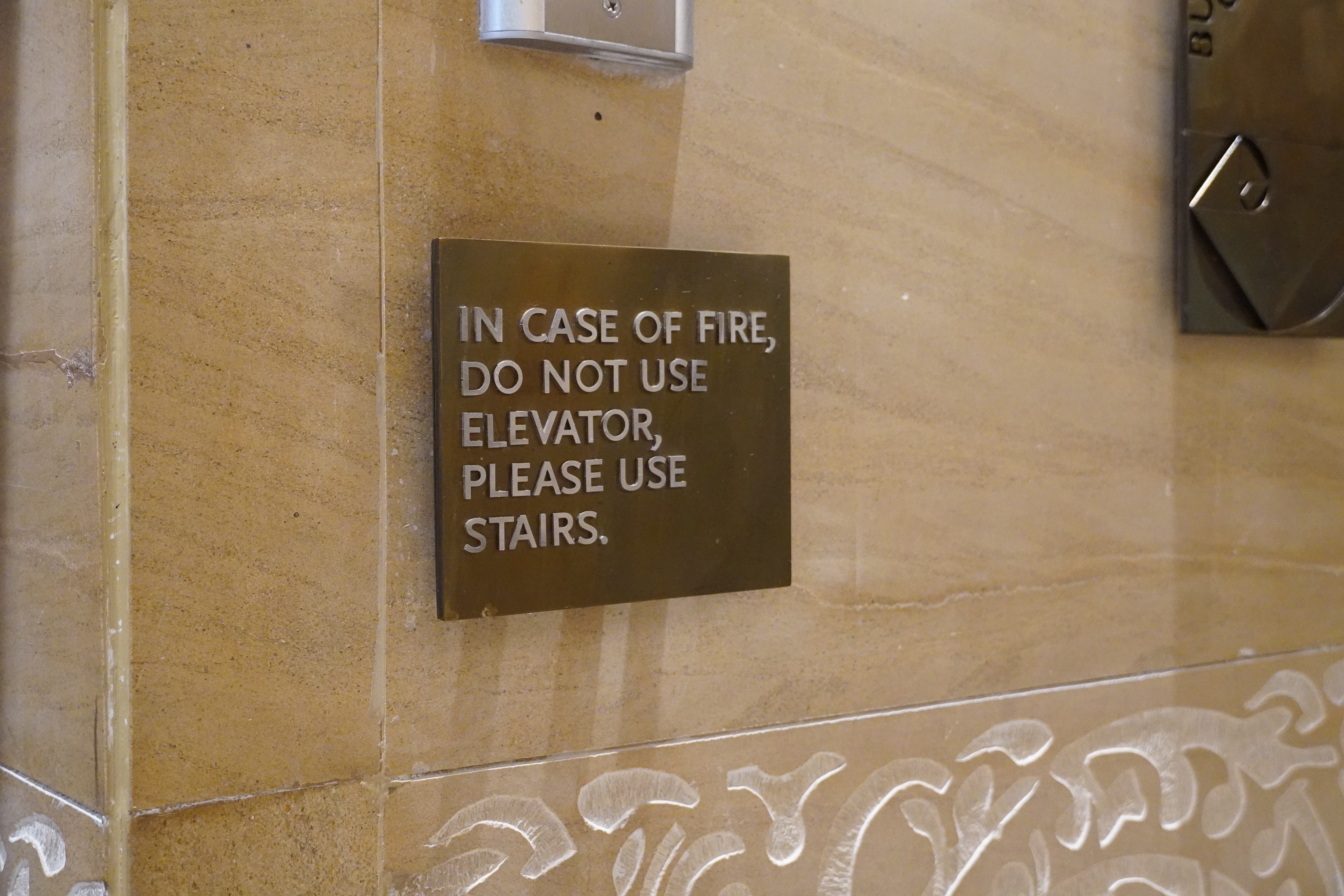 Na slici je natpis na ploči boje bakra koja se nalazi unutar zgrade. Piše "u slučaju požara ne koristite lift nego vas molimo da koristite stepenice ".