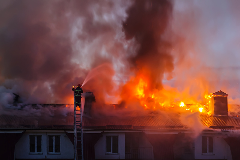 Gasilec stoji na visoki lestvi in gasi gorečo streho stanovanjske stavbe, na kateri se dimniki že podirajo zaradi ognja. 