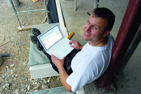 Nasmejani čovek sedi oslonjen na stub građevinskog objekta dok radi na laptopu i gleda u objektiv aparata koji ga slika.