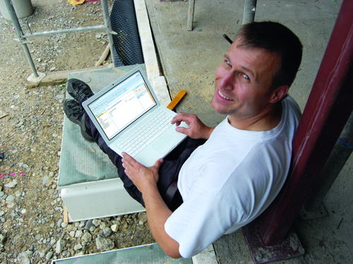Nasmejani čovek sedi oslonjen na stub građevinskog objekta dok radi na laptopu i gleda u objektiv aparata koji ga slika.