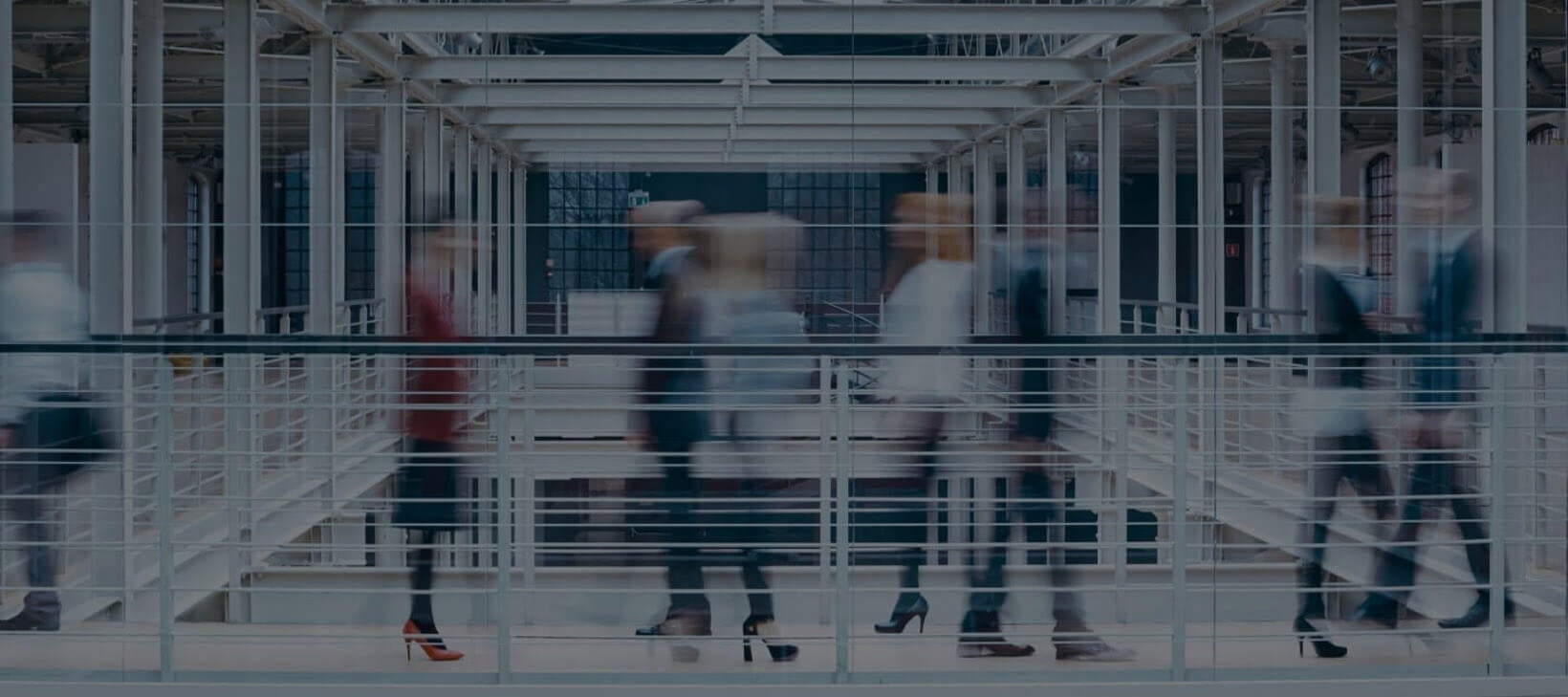 Slika hodnika iz jeklene bele konstrukcije, ki ima jekleno belo ograjo. Na hodniku zamegljene slike sedmih poslovno oblečenih ljudi, ki se premikajo. 