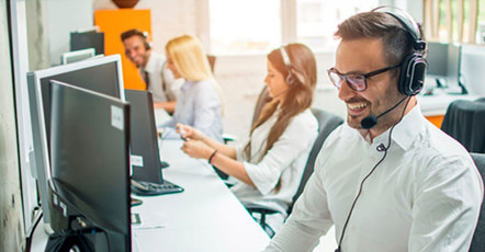 Pet oseb iz tehnične podpore, oblečenih v belo, sedi za temnimi računalniškimi zasloni s slušalkami na glavi in se smeji. 