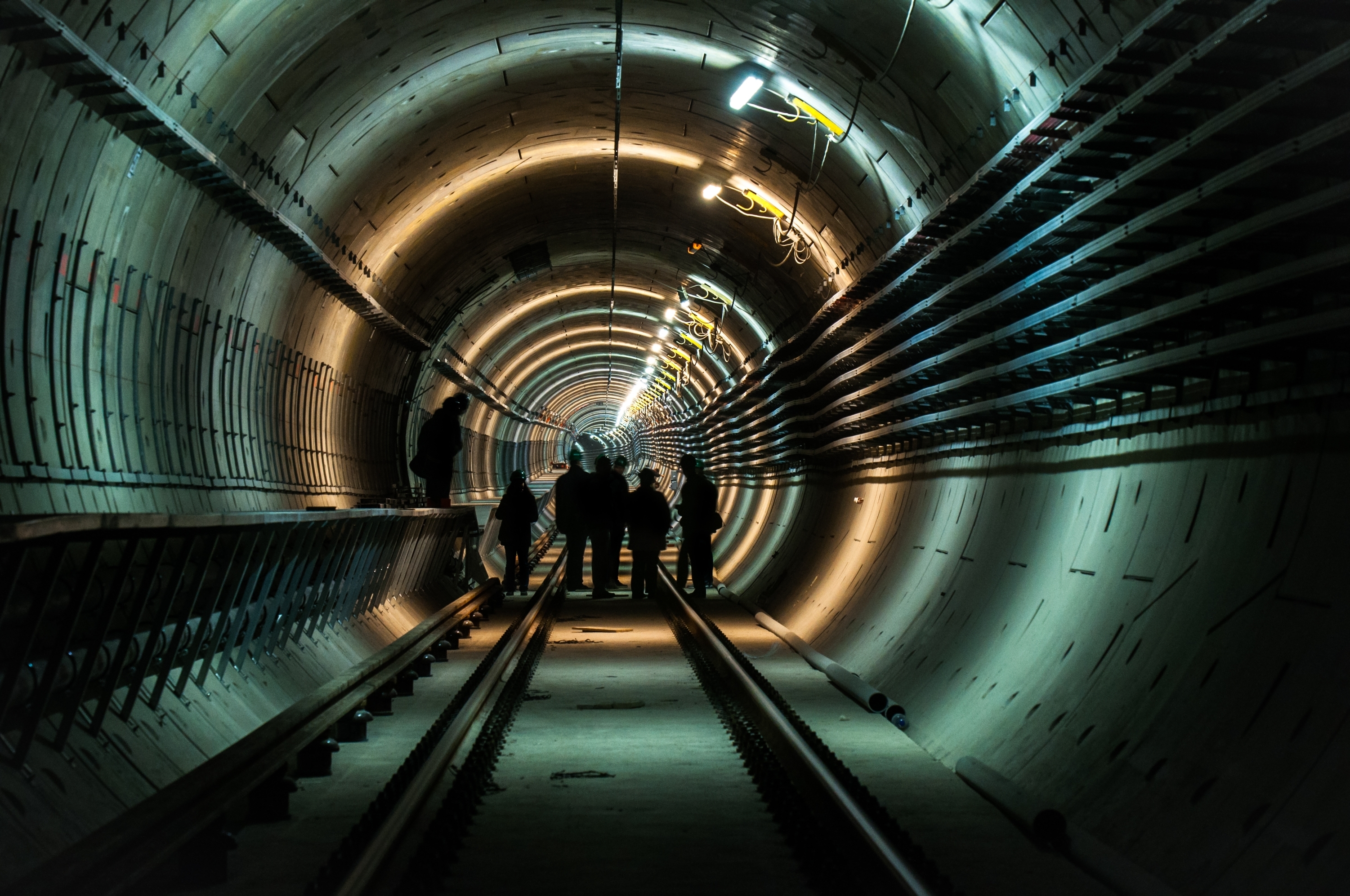 Na slici je unutrašnjost delimično osvetljenog kružnog tunela sa zidnim instalacijama u kome se nalazi nekoliko inspektora.